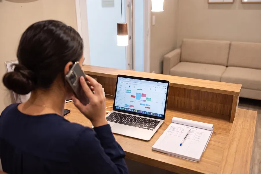 Secretária do consultório ao telefone, com a agenda de consultas aberta no computador